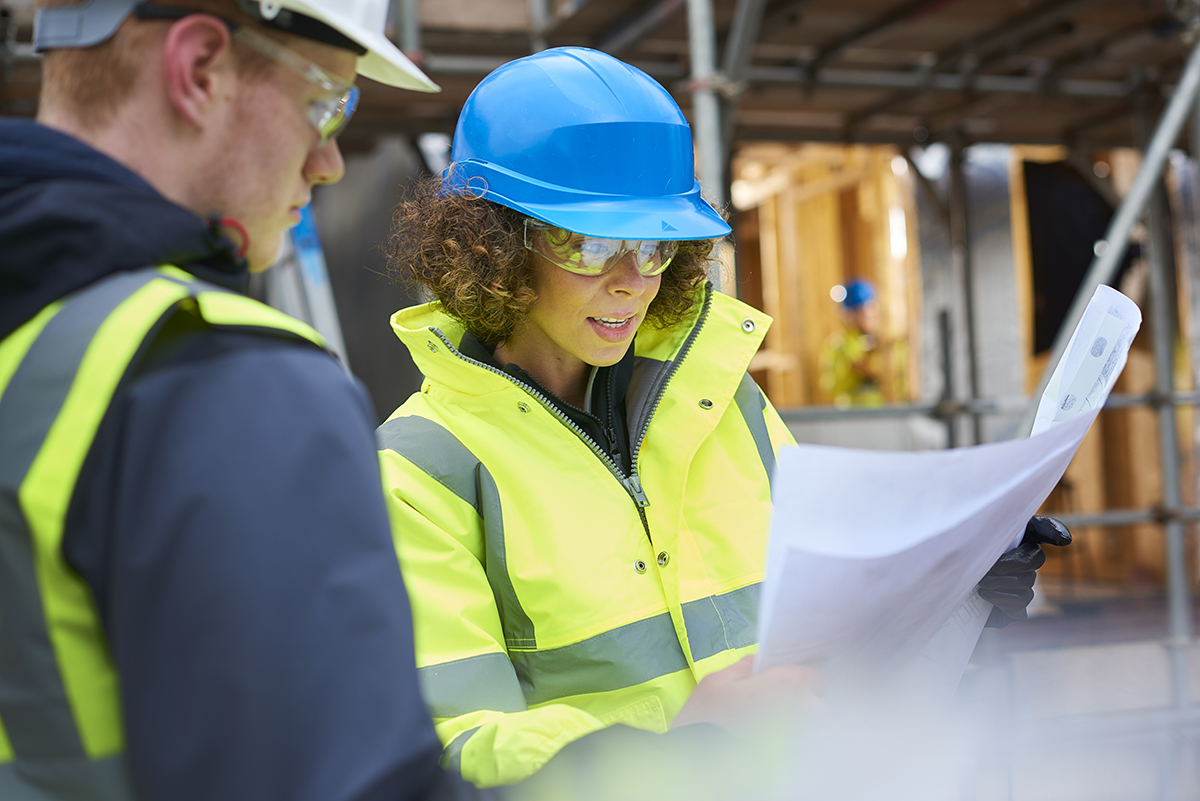 A female construction worker stands on a building site housing development and instructs a co-worker.