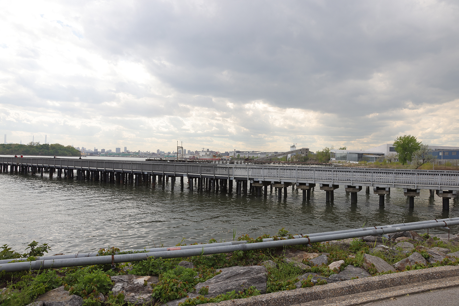 Blue Highway Facility, a long pier in Hunts Point, extends over a body of water, with NYC skyline visible in the distance under a cloudy sky. Green foliage and rocks line the waterfront in the foreground.