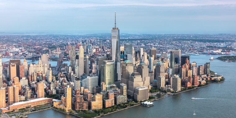 Aerial view of a downtown Manhattan skyline with tall buildings and skyscrapers, surrounded by water, under a cloudy sky.