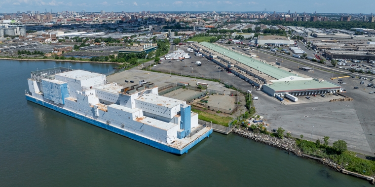 A large white and blue barge is docked at an industrial port area, with warehouses, shipping containers, and city buildings visible in the background, under a clear blue sky.