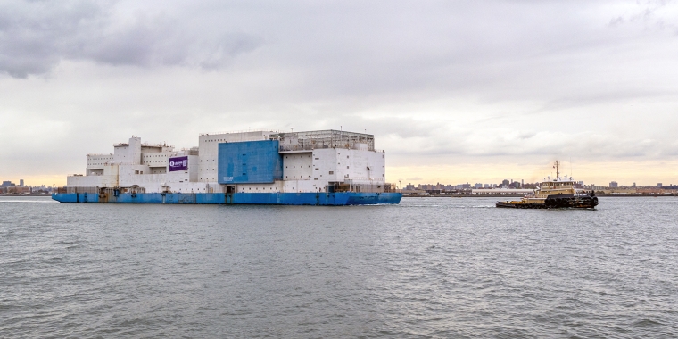 A large white and blue barge, known as the Vernon C. Bain Correctional Center or “the Boat,” sits on the water near a city shoreline, with a smaller tugboat positioned beside it under a cloudy sky.